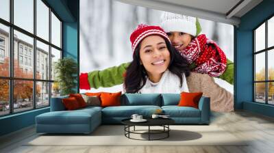 beautiful african american woman piggybacking cheerful daughter and looking at camera in winter park Wall mural