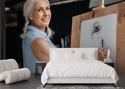 beautiful senior woman smiling at camera while drawing with pencil on easel at art class Wall mural