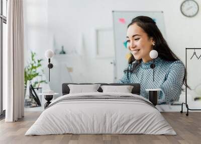 beautiful smiling asian businesswoman sitting at desk and using laptop in office Wall mural