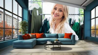 blonde sales assistant in white shirt smiling and holding digital tablet in clothing boutique Wall mural