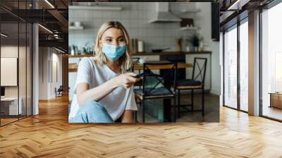 Blonde woman in medical mask holding remote controller at home Wall mural