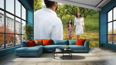 blurred man near happy wife and daughter holding hands near green trees. Wall mural