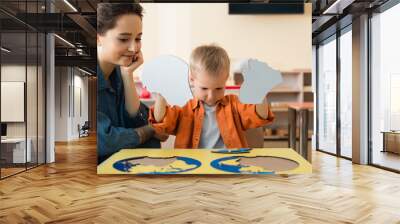 boy holding pieces of puzzle near smiling teacher in montessori school Wall mural