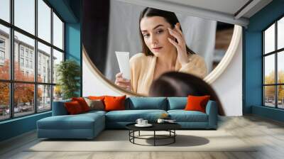 brunette young woman in bathrobe holding tube with cream and looking at mirror in bathroom. Wall mural