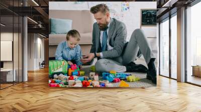 businessman in suit and little son playing with colorful blocks together on floor at home, work and life balance concept Wall mural