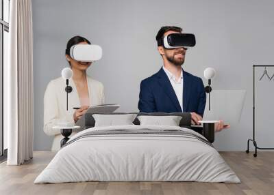 businesswoman holding folder and pen while using vr headsets together with businessman typing on laptop isolated on grey Wall mural