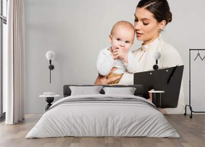 businesswoman with adorable baby on hands holding clipboard with pen, isolated on grey Wall mural