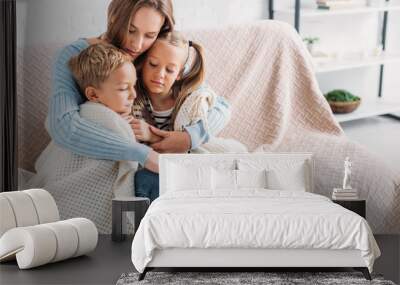 Caring mother wrapping sick children in blanket while sitting on sofa Wall mural