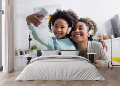 cheerful african american girl in headphones taking selfie on smartphone with happy mother Wall mural