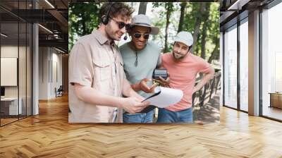 cheerful african american man with vintage camera pointing at clipboard near guide in headset during excursion in city park. Wall mural