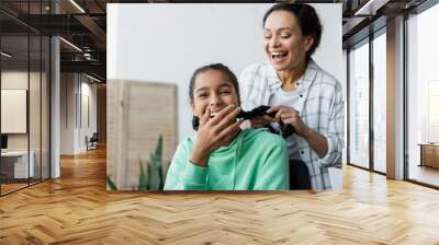 cheerful african american woman braiding hair of laughing daughter covering mouth with hand Wall mural