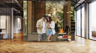 Cheerful and stylish couple holding wine near grill and smoke with vacation house at background Wall mural