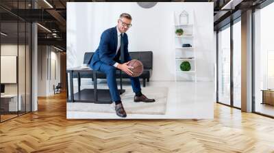 cheerful businessman in suit holding basketball in hands while sitting on coffee table Wall mural