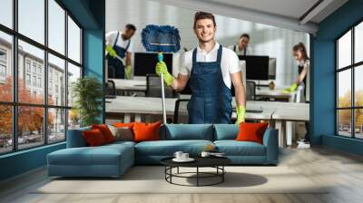 cheerful cleaner holding mop while looking at camera near multicultural colleagues Wall mural