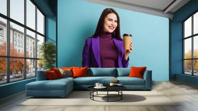 cheerful girl looking at camera while holding coffee to go on blue background Wall mural