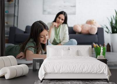 cheerful girl lying on carpet and drawing near blurred nanny sitting on sofa. Wall mural