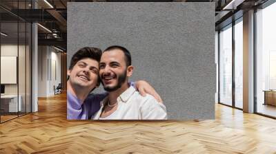 Cheerful homosexual couple in casual clothes taking selfie on smartphone together while standing near grey building wall on urban street at daytime, banner Wall mural