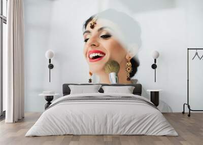 cheerful indian bride applying face powder with cosmetic brush on white Wall mural