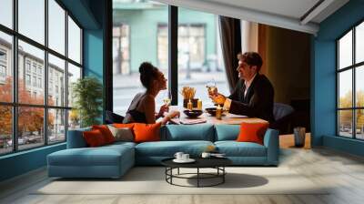 cheerful interracial couple in elegant attire holding glasses with wine during date in restaurant Wall mural