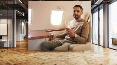 cheerful man holding champagne glass while sitting in first class of private jet Wall mural