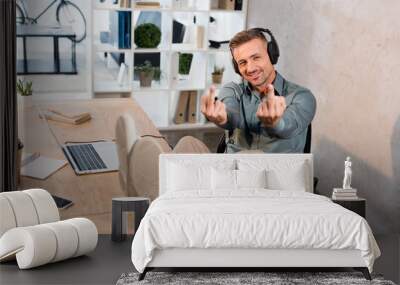 cheerful man listening music in headphones and showing middle fingers in office Wall mural