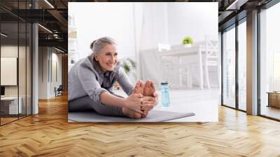 cheerful mature woman with grey hair stretching on yoga mat near sports bottle Wall mural