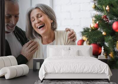 cheerful middle aged woman laughing while holding bauble near husband and christmas tree, banner Wall mural