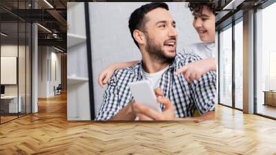 cheerful muslim boy pointing at smartphone in hands of smiling father, banner Wall mural