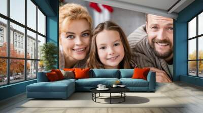 Cheerful parents looking at camera near daughter during new year celebration at home, banner Wall mural