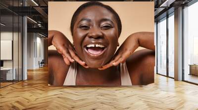 cheerful plus size african american woman smiling with hands near her face on beige background Wall mural