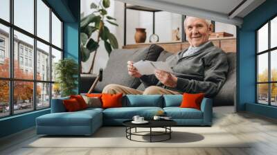 cheerful senior man with grey hair holding photos and sitting on sofa Wall mural