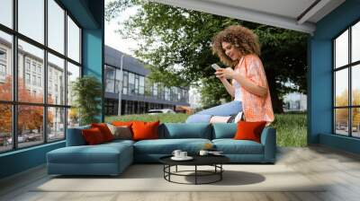 cheerful woman with curly hair using smartphone while sitting on lawn in green park. Wall mural