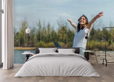 cheerful woman with outstretched hands sitting with laptop near lake Wall mural