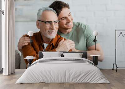 cheerful young man with closed eyes hugging bearded and mature father in eyeglasses Wall mural