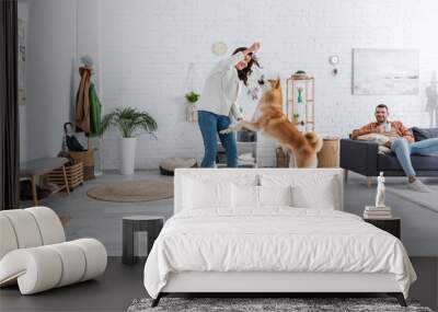 cheerful young woman playing with akita inu dog near bearded boyfriend with cup of coffee sitting on couch. Wall mural