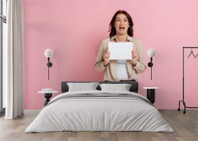 Confused woman looking at camera while holding empty card on pink background, concept of body positive Wall mural