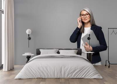 corporate asian businesswoman with grey hair talking on smartphone and holding laptop isolated on grey Wall mural