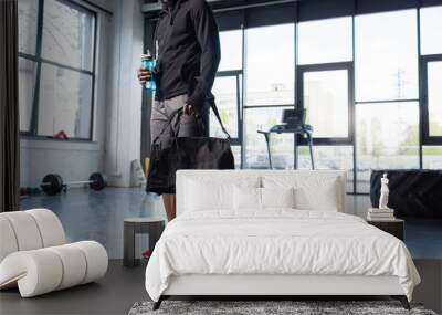 cropped shot of african american man in sportswear holding bag and bottle of water in gym Wall mural