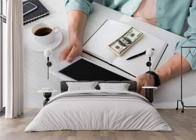 Cropped view of freelancer holding digital tablet near coffee and dollar banknotes on notebook at table, concept of earning online Wall mural