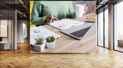 Cropped view of teleworker holding cup and pen near notebook, papers and devices on blurred foreground Wall mural