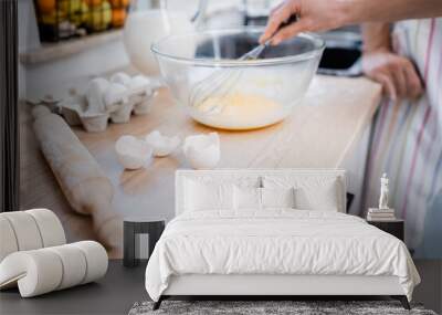 Cropped view of woman mixing eggs in blurred bowl near rolling pin on worktop in kitchen. Wall mural