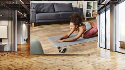 Curly African American woman in active wear gracefully holding a yoga pose on a yoga mat at home. Wall mural