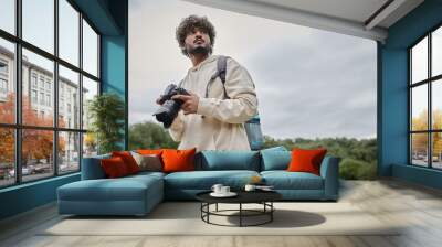 curly indian photographer in hoodie holding professional camera, travel and adventure concept Wall mural