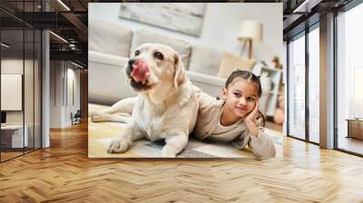 cute elementary age girl in casual attire lying on carpet with labrador in modern living room Wall mural