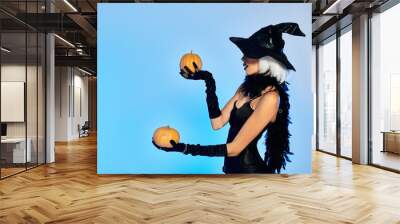 Dressed in a striking witch costume, a young woman elegantly balances two pumpkins while posing. Wall mural