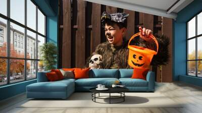 excited asian boy in werewolf costume holding halloween bucket and spooky skull while looking away Wall mural