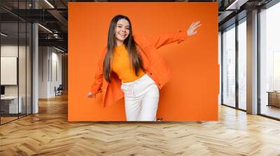 Fashionable and cheerful brunette teenage girl in denim jacket and pants looking at camera while posing isolated on orange, cool and confident teenage girl, gen z fashion Wall mural