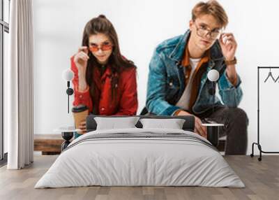 fashionable hipster couple with coffee cups adjusting eyeglasses and sitting on bench isolated on white Wall mural