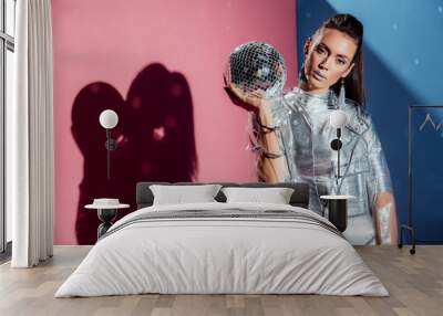 fashionable model in silver bodysuit and raincoat posing with disco ball on pink and blue background Wall mural