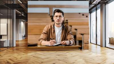 focused male student in glasses sitting at desk, looking at camera while writing in notebook during lesson in classroom Wall mural